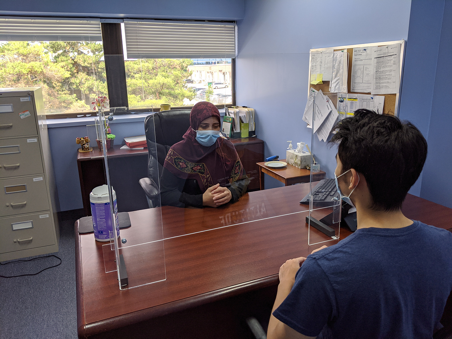 A settlement worker seeing a client in her office, they're both wearing masks over their mouths with glass partition between them.